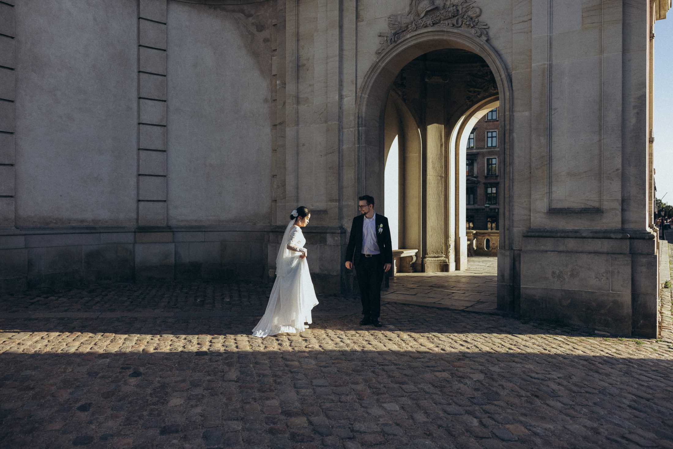Bride and groom beneath a grand stone archway, sunlight falling on the cobblestones