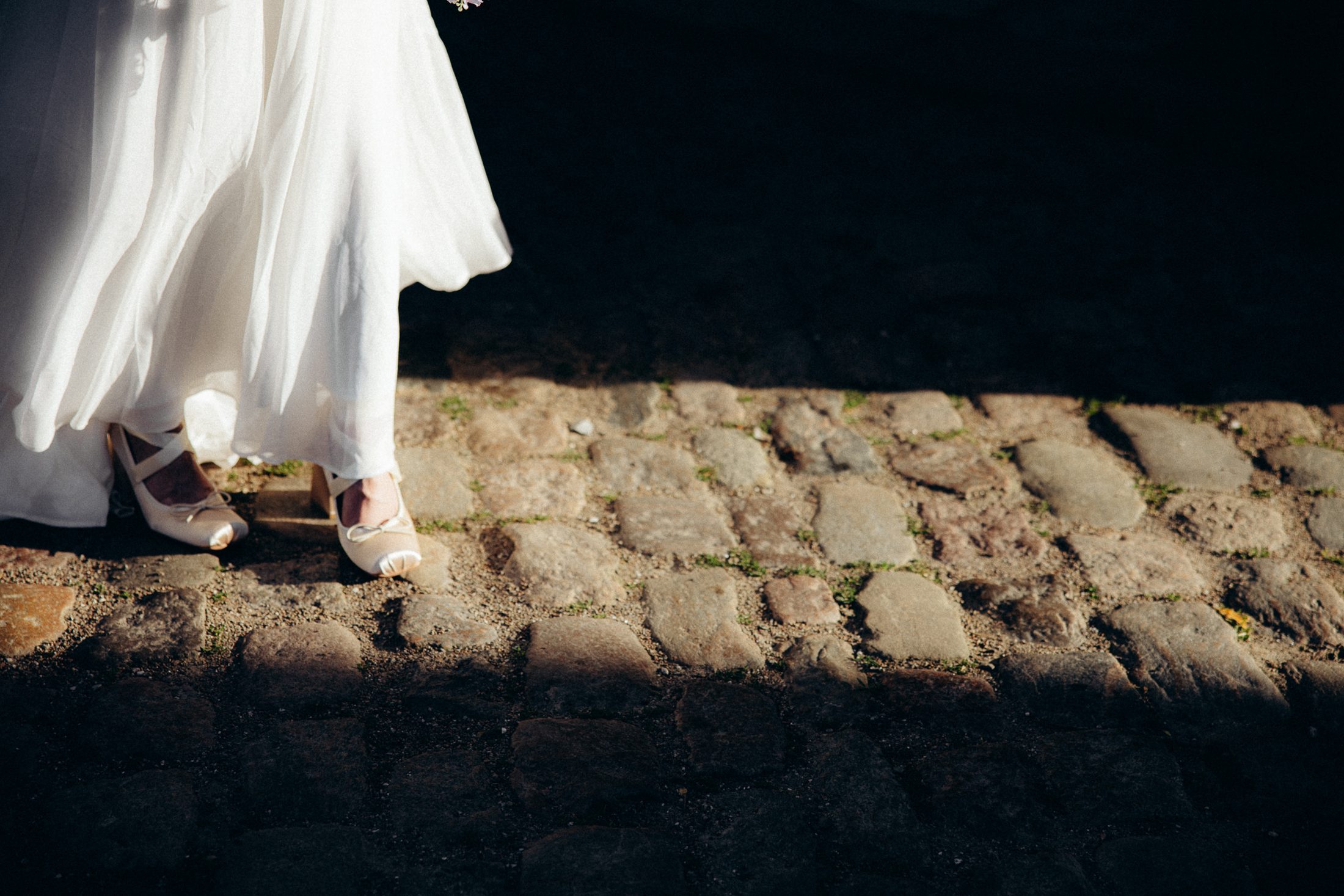 The bride's shoes and dress hem in a strip of sun on the cobblestones